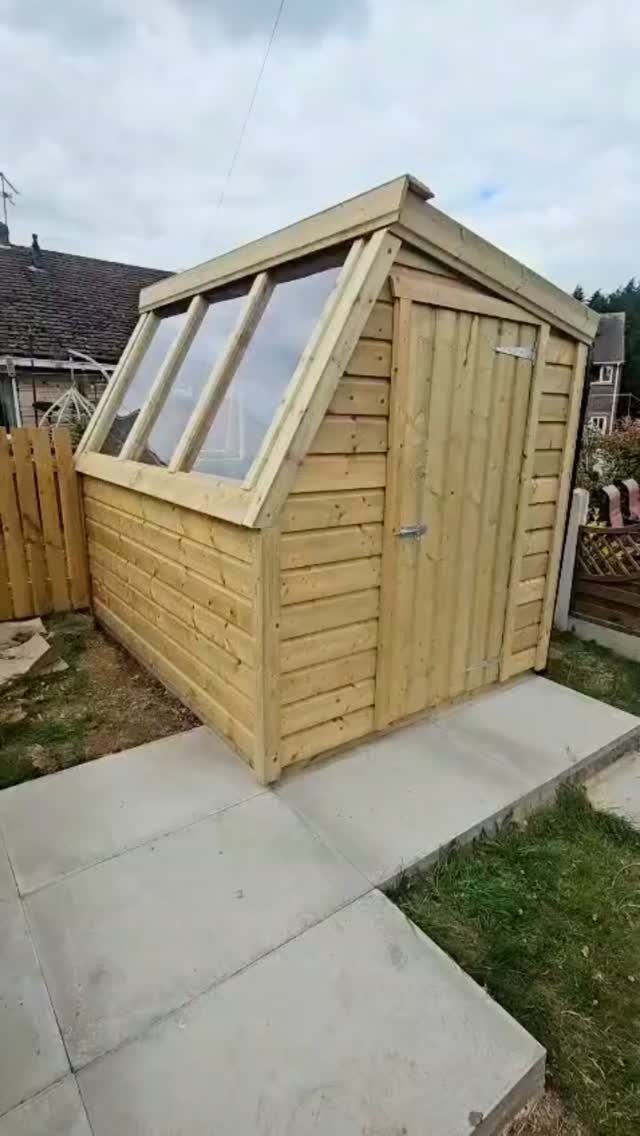 A small wooden garden shed with a large glass window sits on a concrete patio.