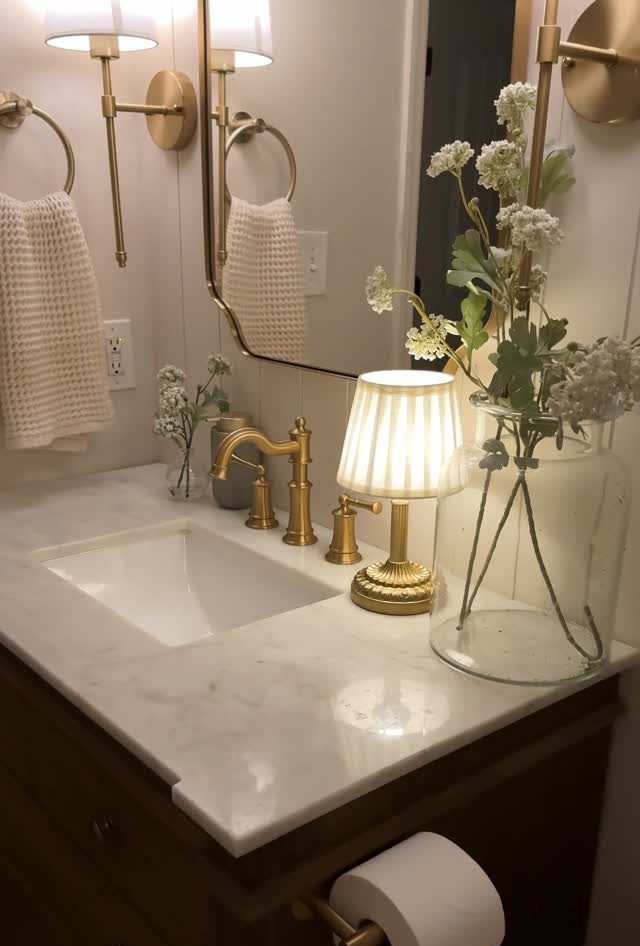 Gold bathroom vanity featuring a marble countertop, gold faucet, and elegant pleated lamp with white flowers.