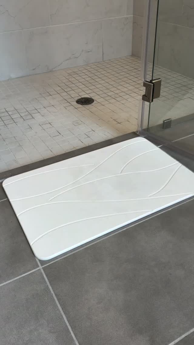 A white textured stone bath mat sits on grey floor tiles outside a glass shower.