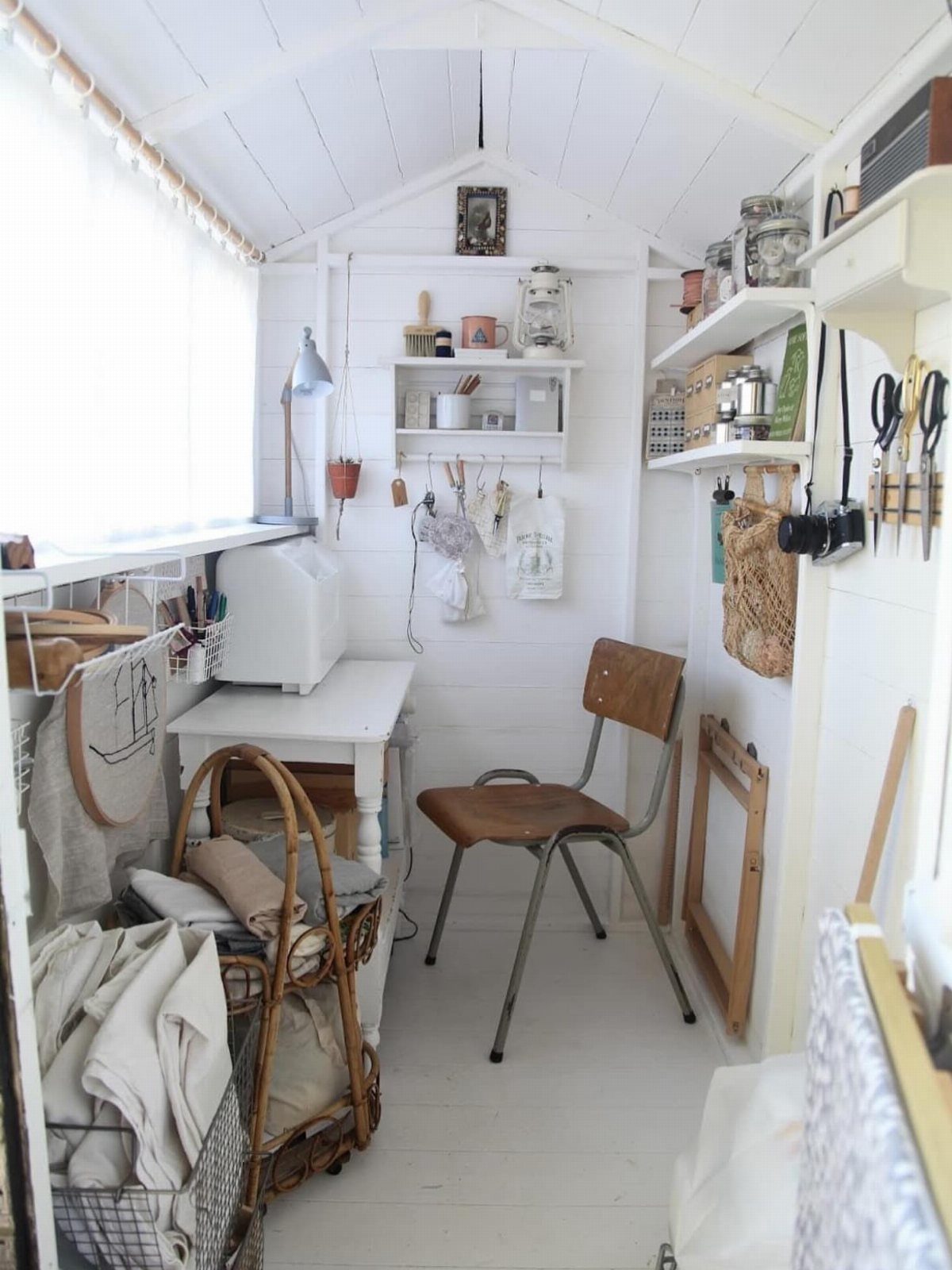 Interior view of a white wooden art studio shed with a desk, chair, and art supplies.