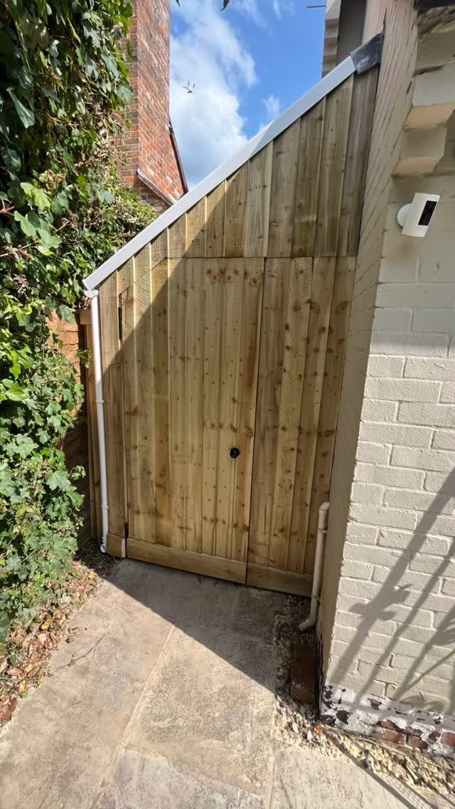 A wooden garden shed with a slanted roof tucked between a brick wall and green foliage.