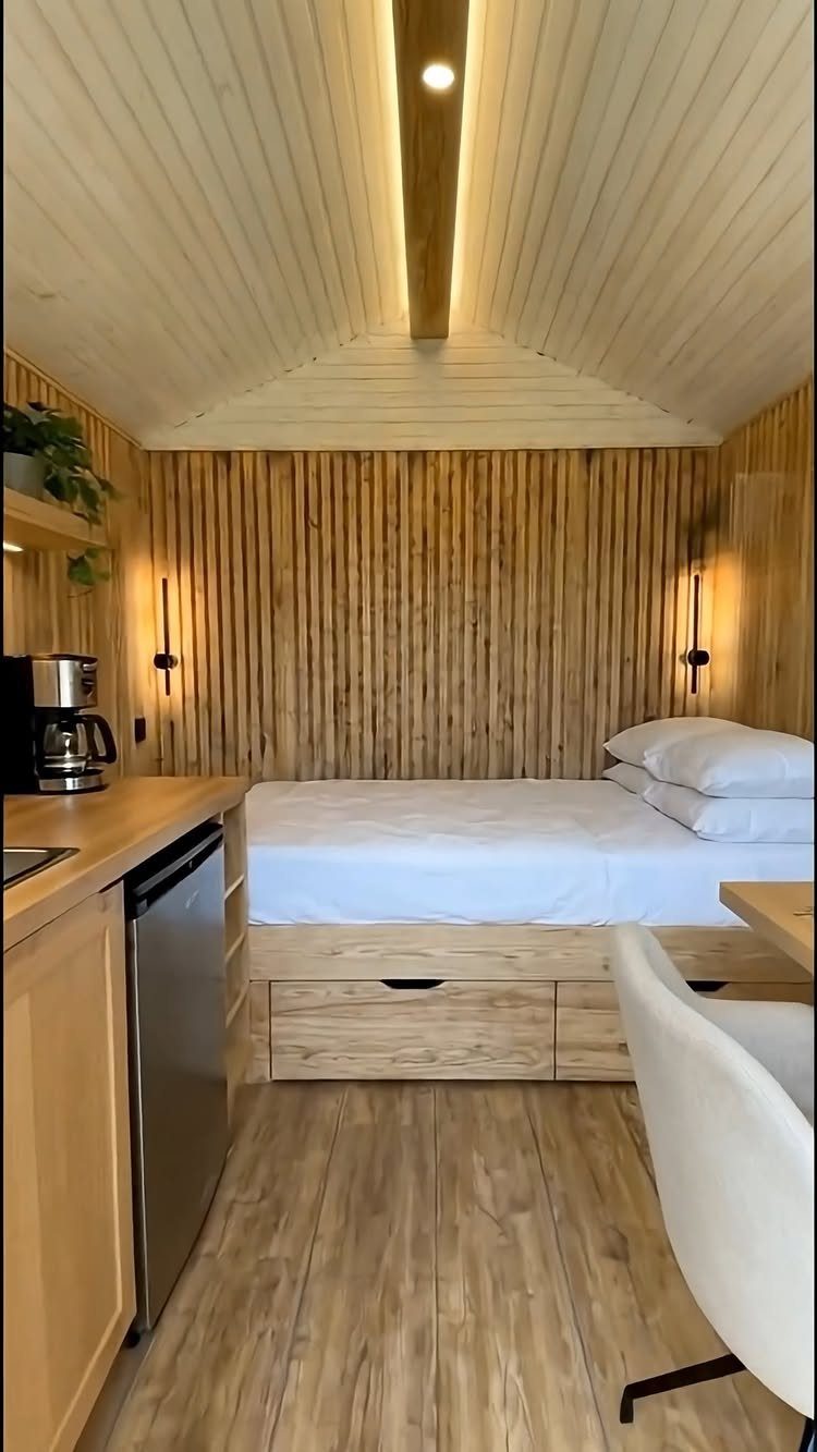 Interior of a modern wooden tiny house featuring a bed, kitchenette, and light wood paneling.