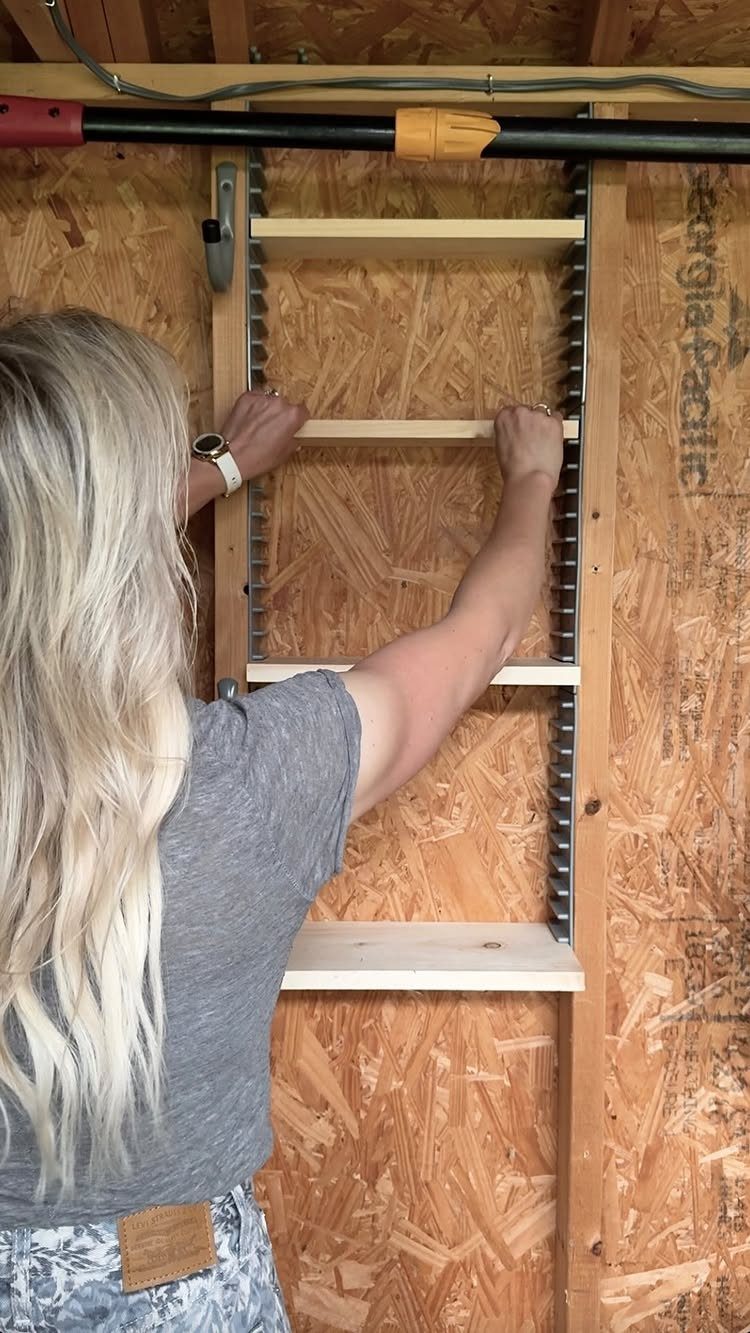 A woman installing wooden shelves onto a metal adjustable track system against a wooden wall.