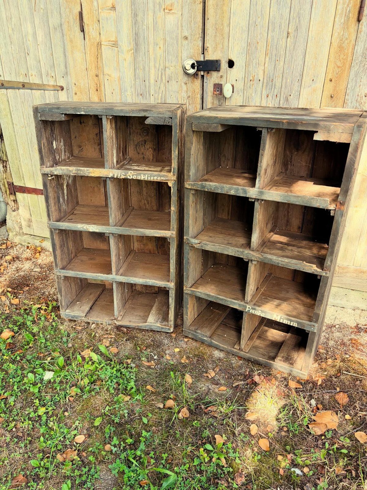 Two rustic wooden cubby shelving units sitting on grass in front of a wooden shed.