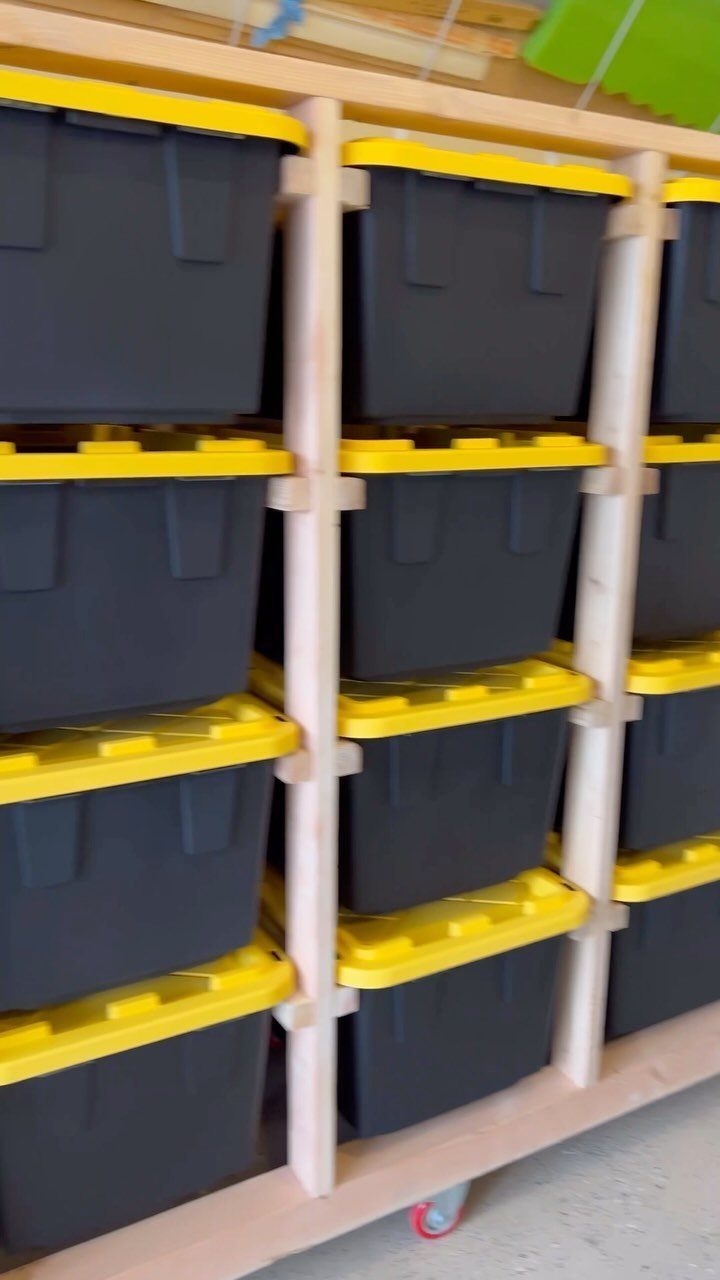 Wooden rolling shelves holding multiple black storage bins with yellow lids on red wheels.