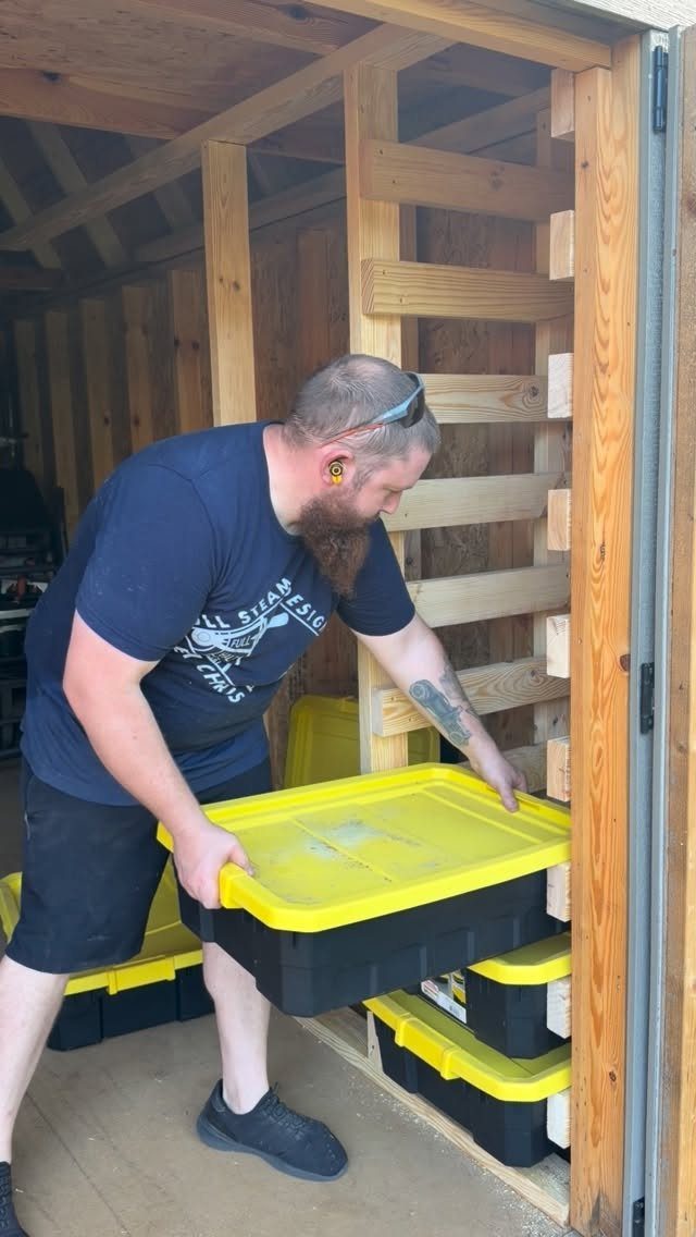 Man placing a yellow plastic storage bin onto a wooden slatted wall shelving system inside a shed.