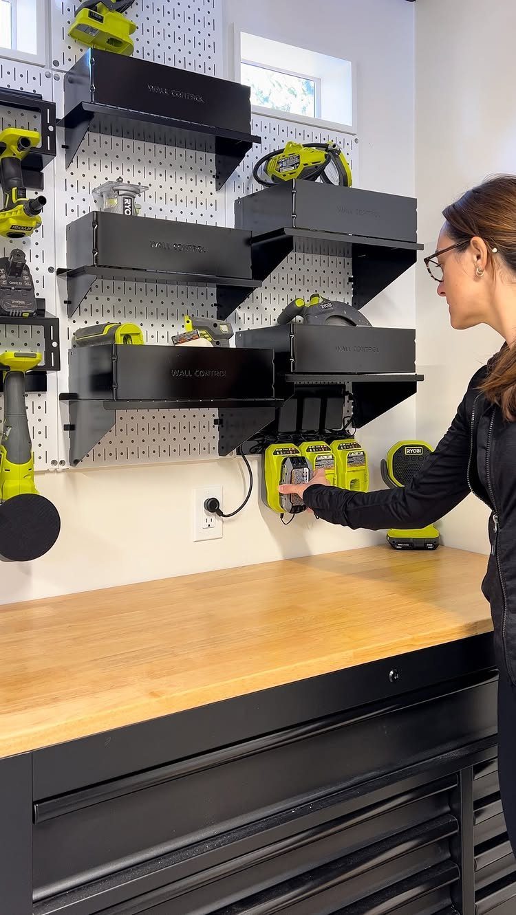A woman organizes yellow power tools on a white pegboard with black wall-mounted storage bins.