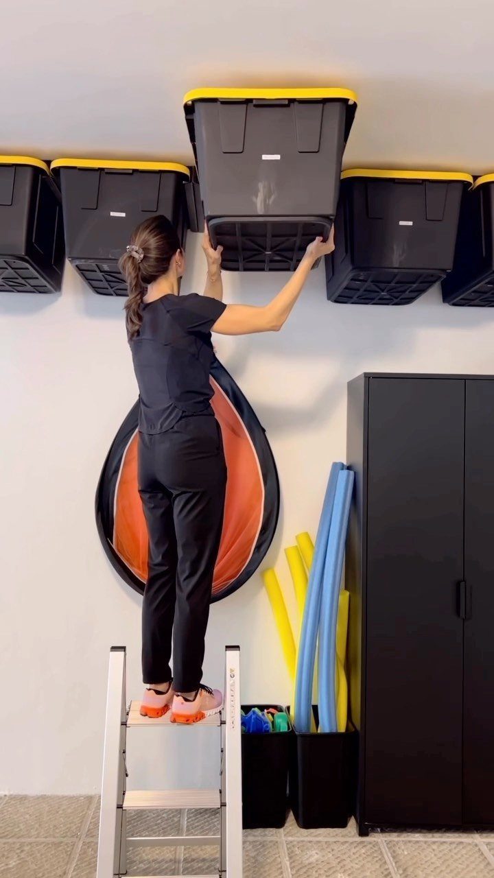 A woman on a ladder organizing black and yellow storage bins on a ceiling rack.