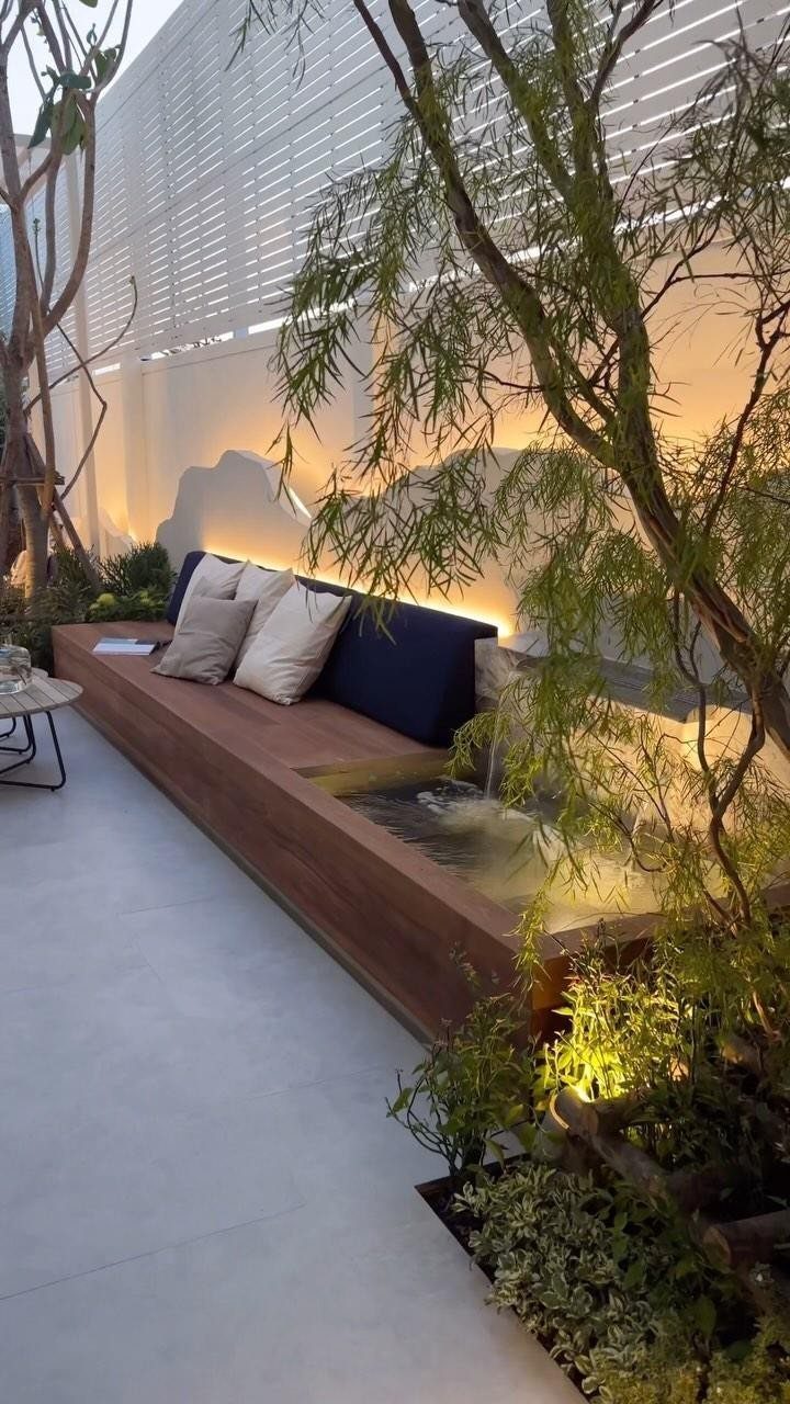 A modern wooden built-in bench with cushions next to a small water feature and greenery.