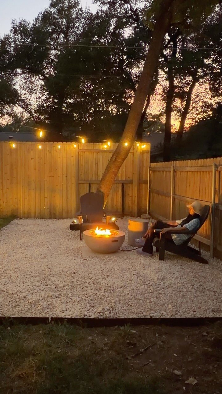 A person relaxing in a chair beside a lit fire pit in a gravel backyard.