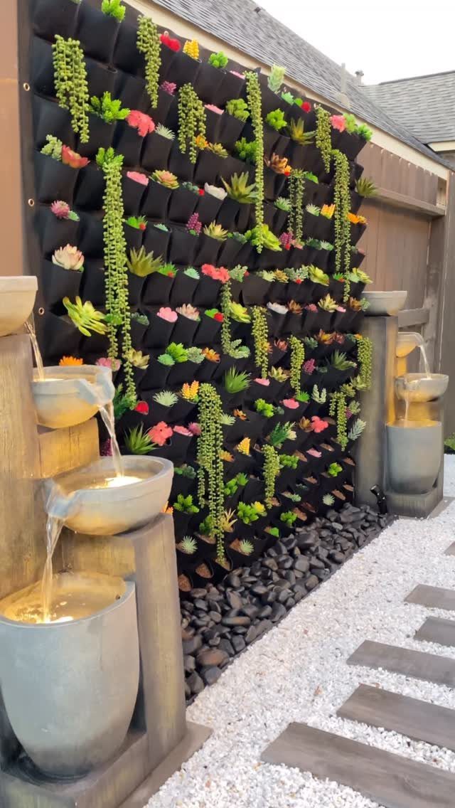 A black vertical wall-mounted planter filled with colorful succulents next to stone water fountains.