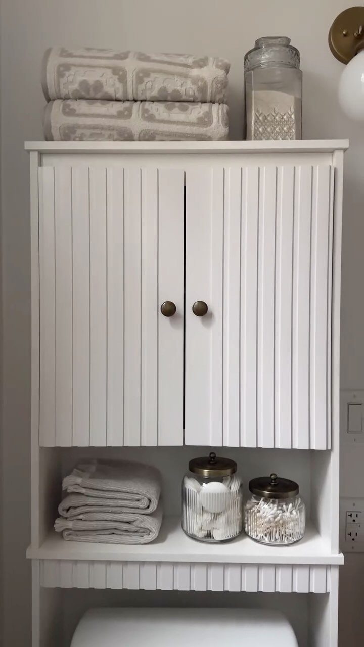 White wooden over-the-toilet storage unit with towels and glass jars on shelves.