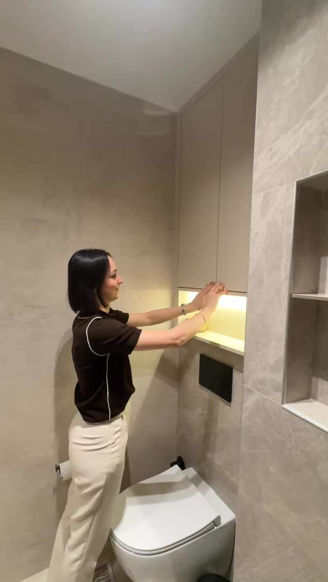 Woman reaching into a glowing recessed wall niche in a modern grey tiled bathroom.