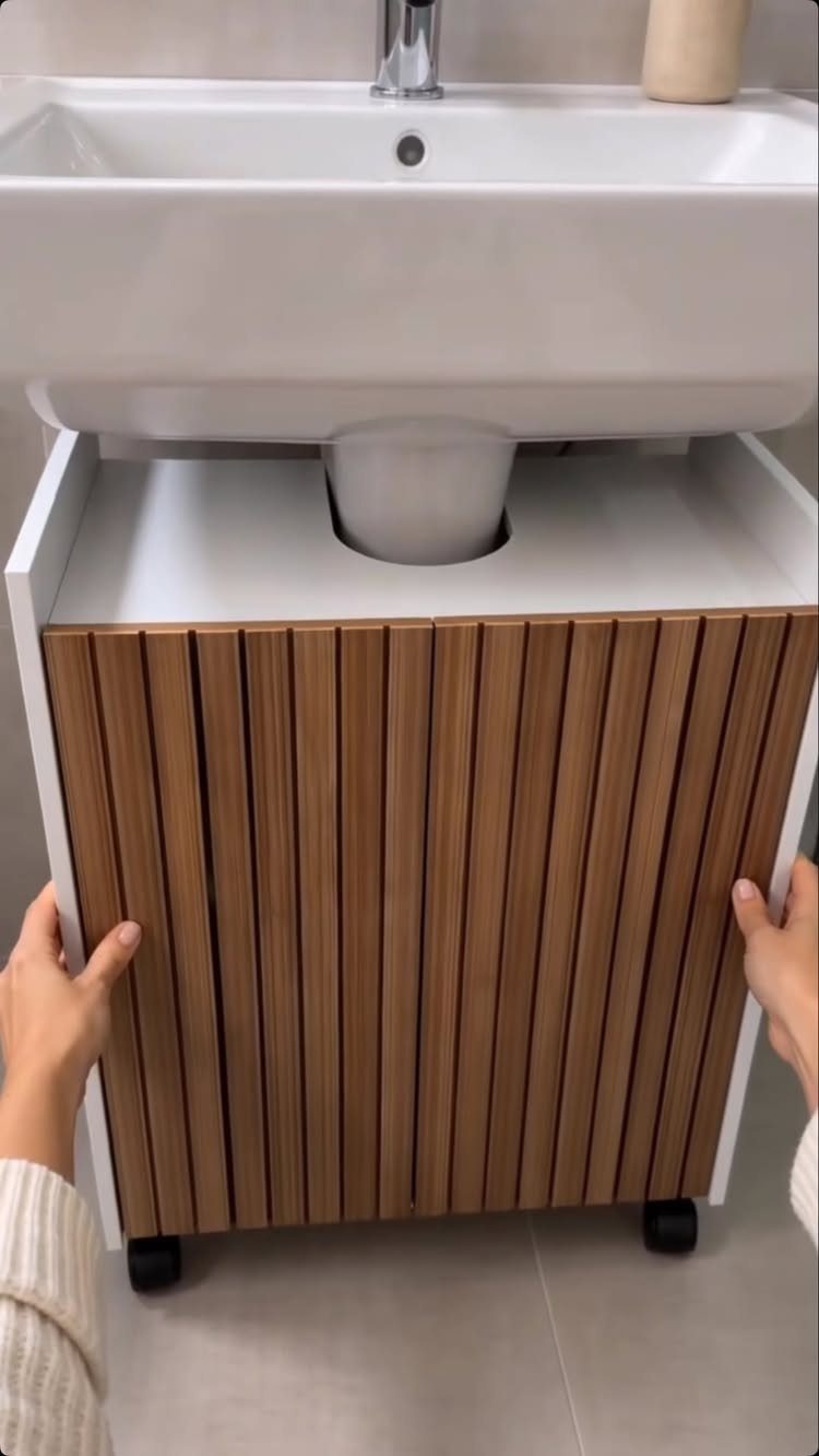 Hands pulling a wooden slatted rolling storage unit from under a white bathroom sink.