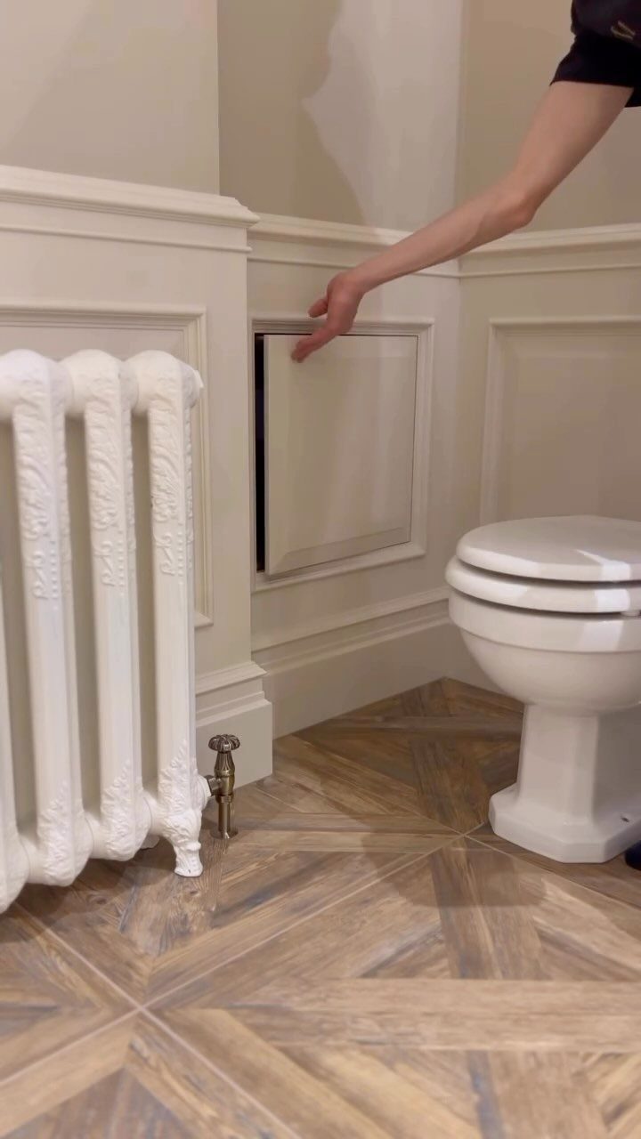 A hand opening a small secret cabinet built into white bathroom wainscoting.