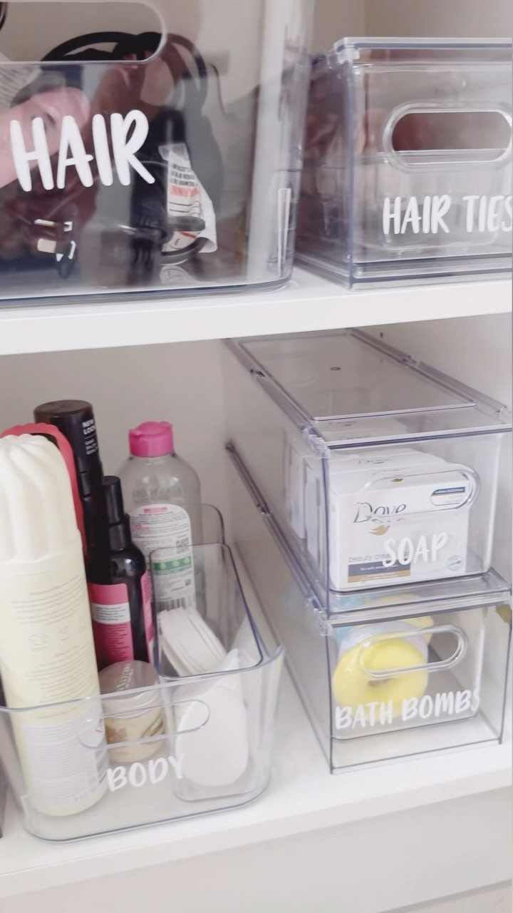 Clear plastic storage bins with white text labels containing hair products and toiletries on white shelves.