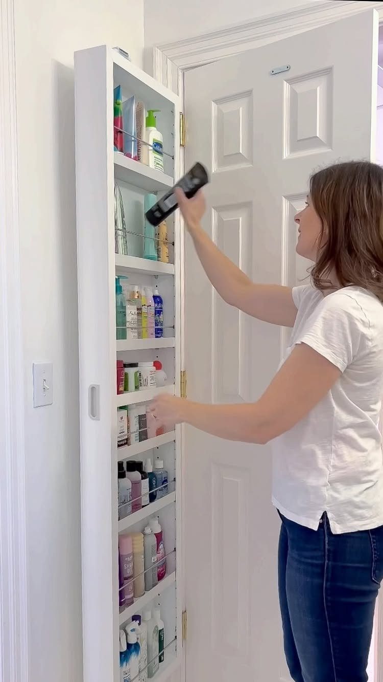 A woman organizing various colorful toiletry bottles inside a slim white storage cabinet mounted on a door.