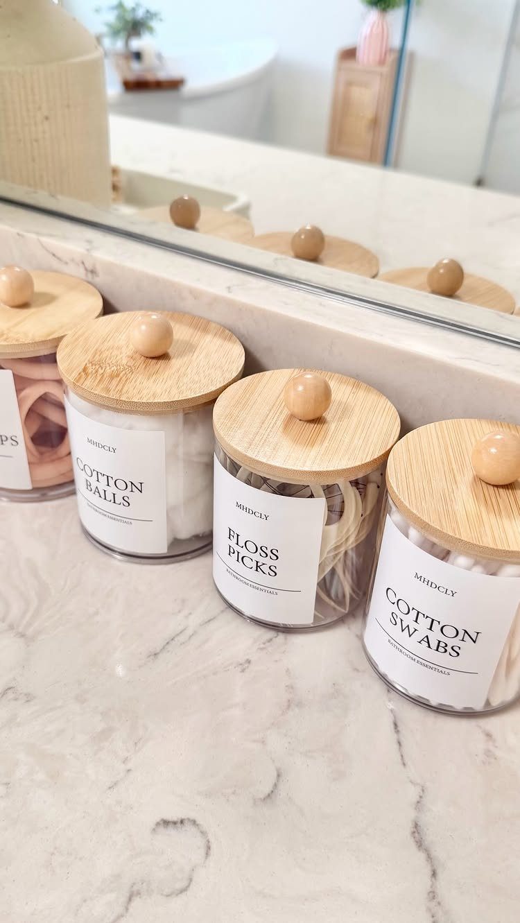 Four glass canisters with wooden lids containing bathroom essentials on a marble countertop.