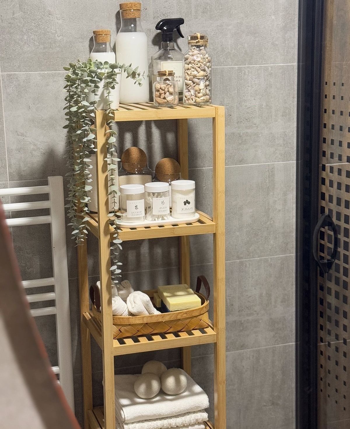 A bamboo four-tier shelf holding bathroom essentials against a grey tiled wall.