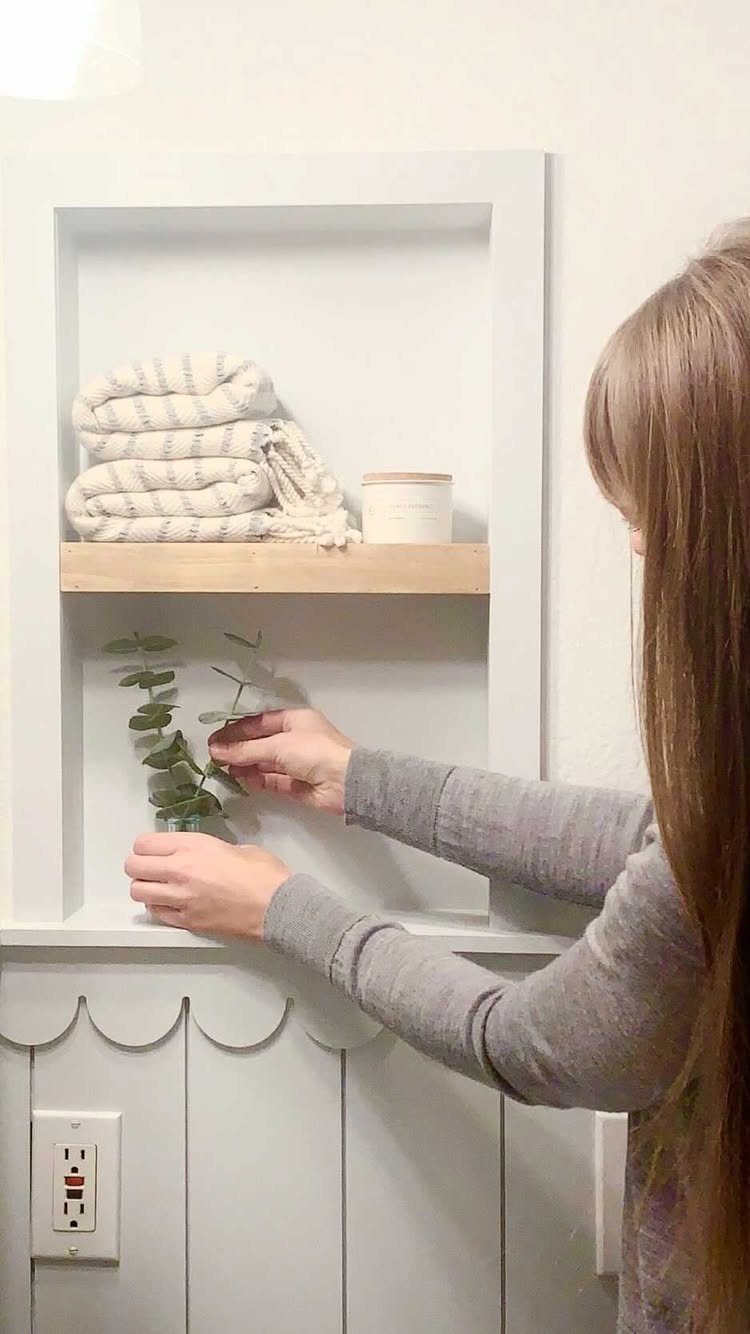 Hands reaching toward a recessed wall niche with a wooden shelf holding folded white towels.