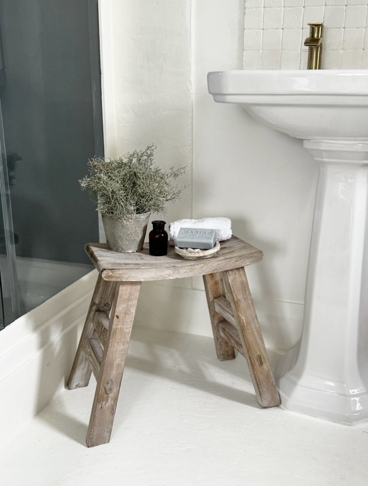 A rustic wooden stool serving as a side table next to a white pedestal sink.