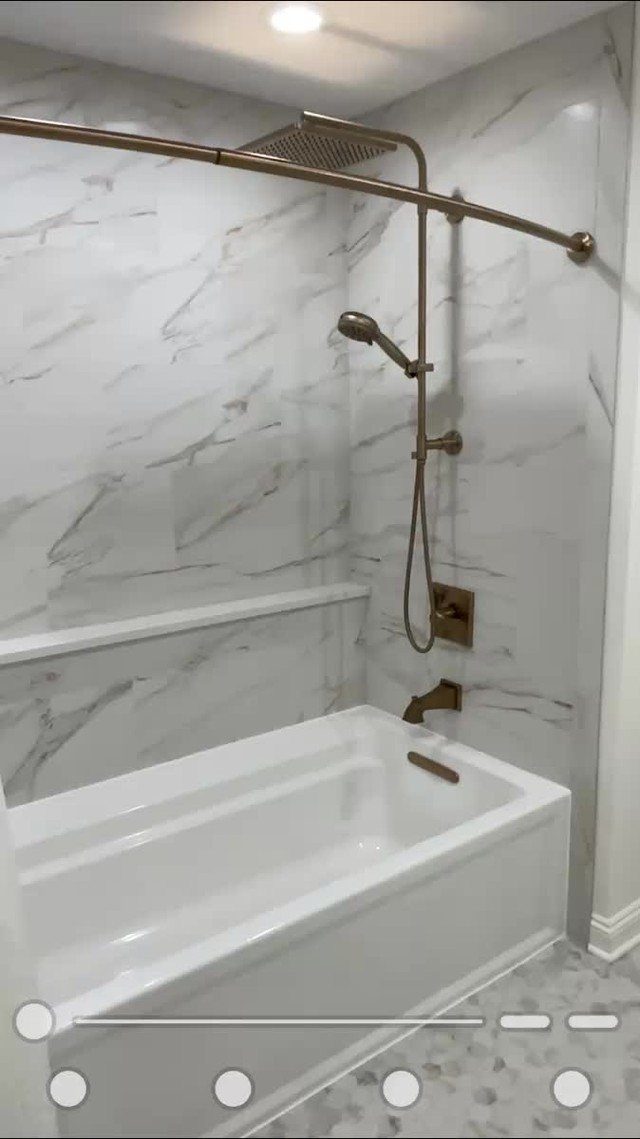 A white bathtub with bronze shower fixtures against marble patterned walls.