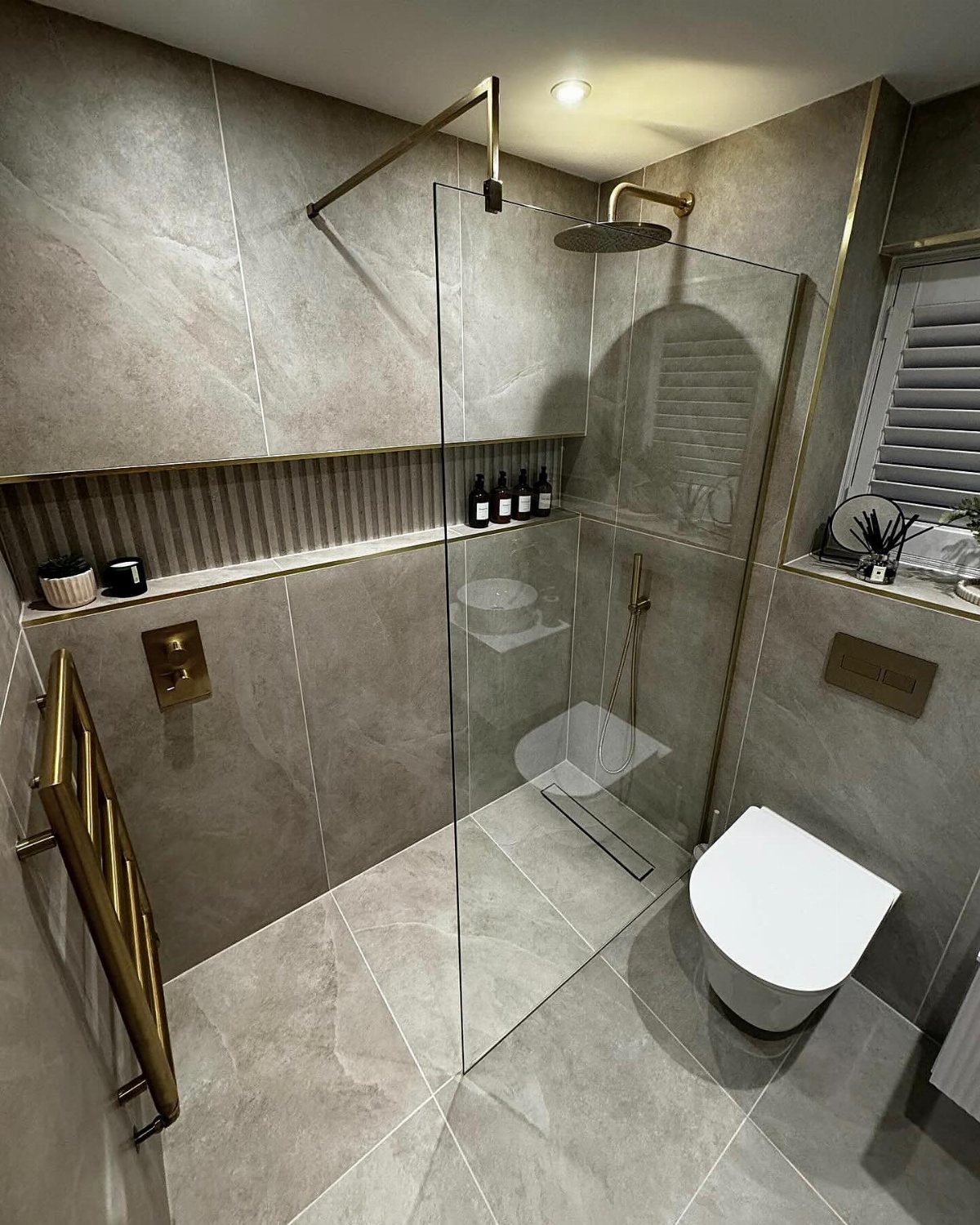 Modern grey tiled bathroom with a glass walk-in shower and gold fixtures.