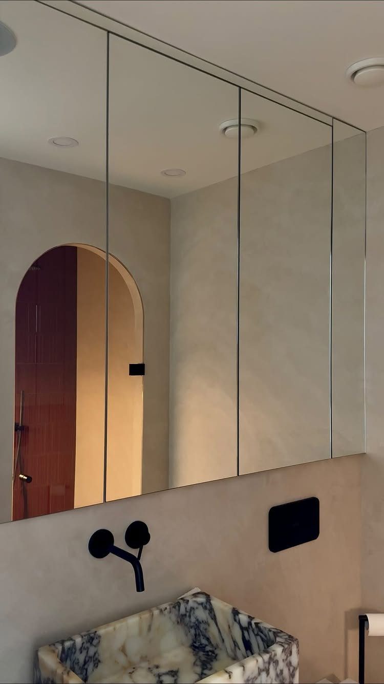 Four large mirrored cabinet panels mounted above a marble sink and black faucet.
