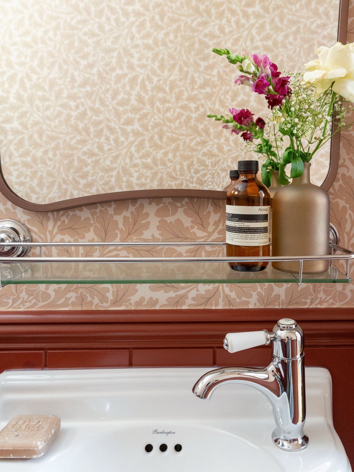 White sink with chrome faucet, glass shelf, and floral arrangement against patterned wallpaper.