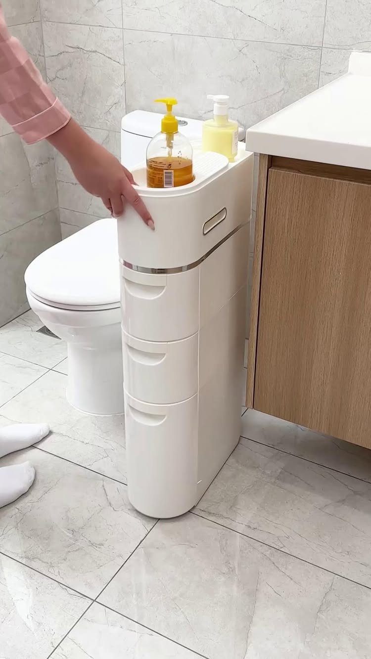 A white slim rolling storage cart positioned between a toilet and a wooden bathroom vanity.