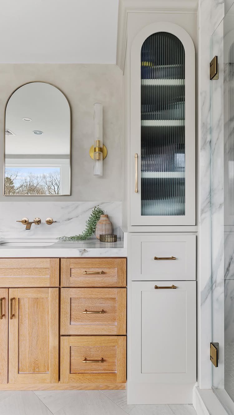 Light wood bathroom vanity with marble countertop and tall white fluted glass cabinet.
