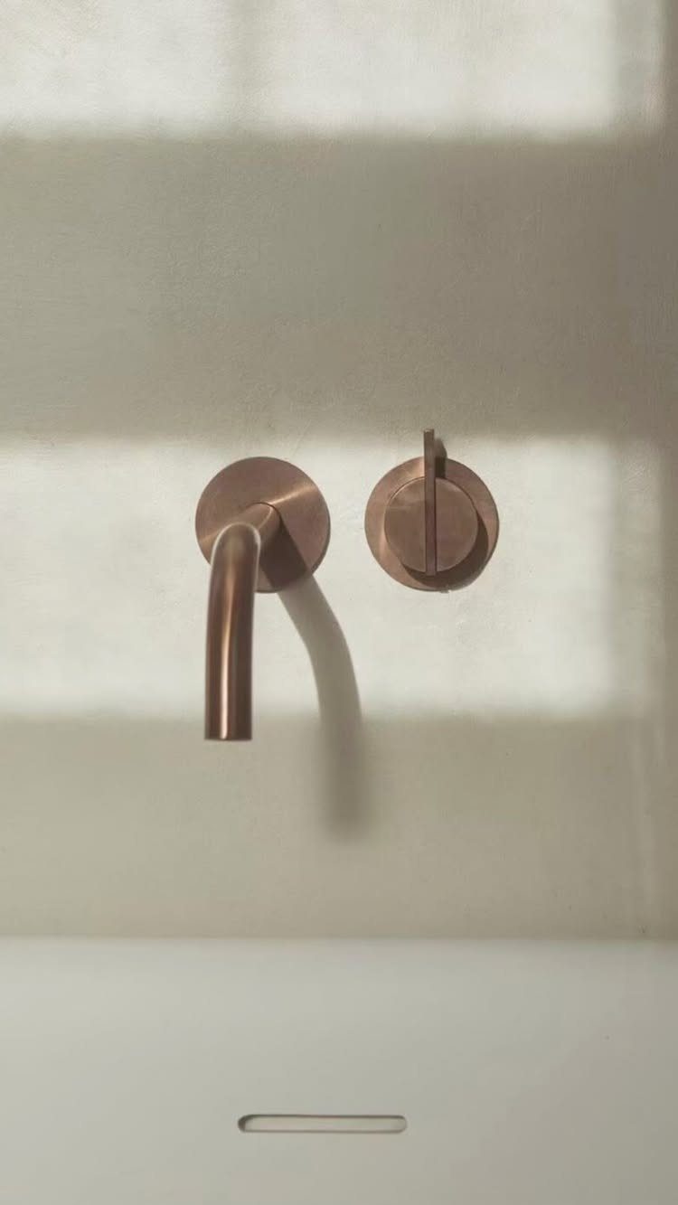 Copper wall-mounted faucet and circular control handle mounted on a light wall above a white sink.