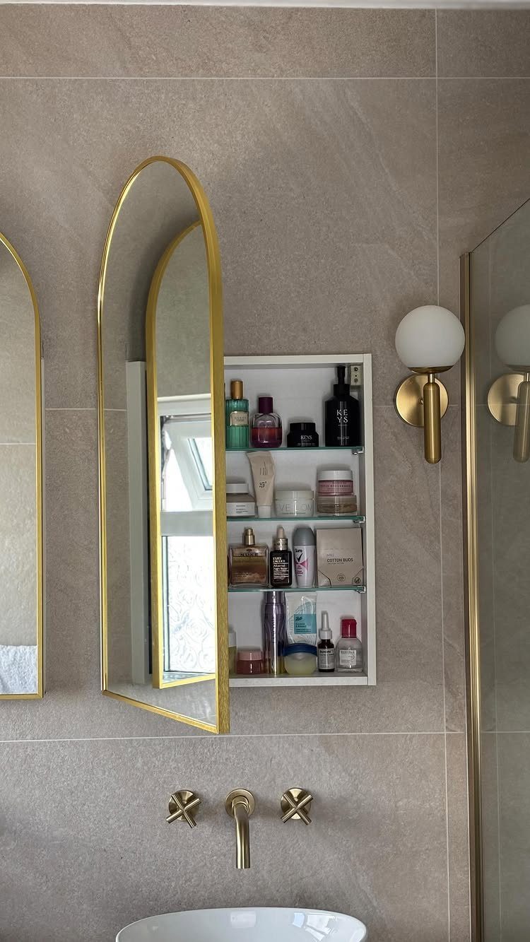 An open arched gold-framed medicine cabinet filled with various skincare products on a tiled wall.
