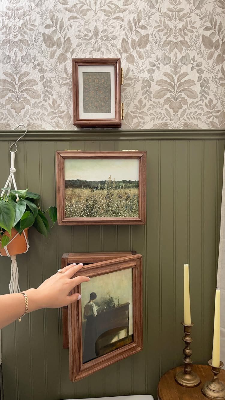 A hand opens a wooden picture frame to reveal a hidden medicine cabinet on a wall.