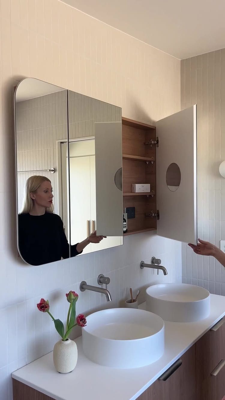 A woman opens a white surface-mounted medicine cabinet with a wooden interior above bathroom sinks.