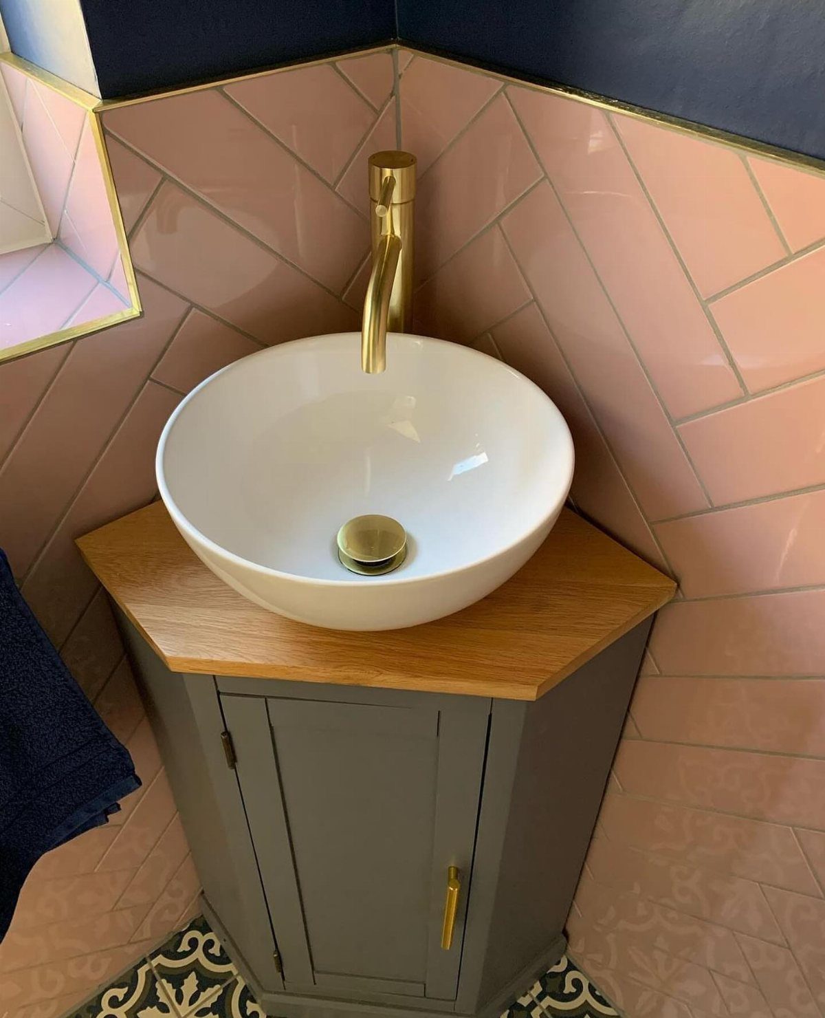 A white vessel sink with gold hardware sits on a wooden corner vanity cabinet.