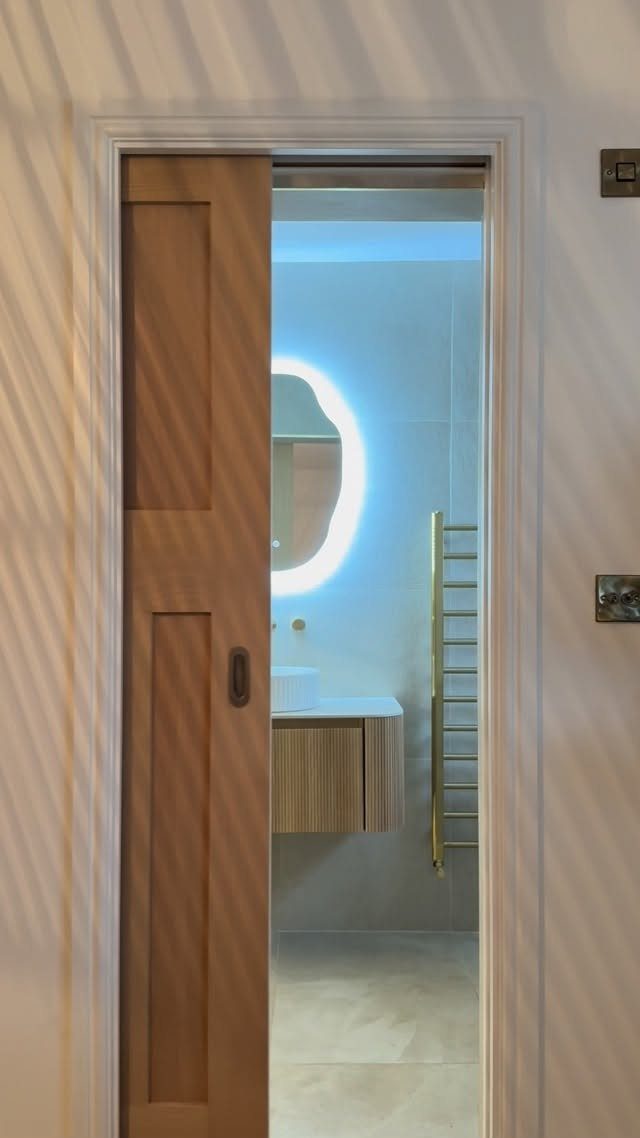A wooden pocket door partially open, revealing a modern bathroom with a floating vanity and backlit mirror.