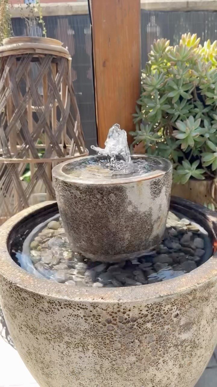 A stone water fountain with bubbling water and dark river stones in a garden setting.