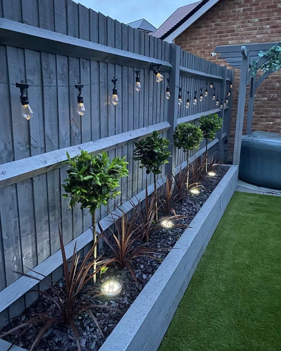 String lights with Edison bulbs hanging along a grey wooden fence above a garden bed.