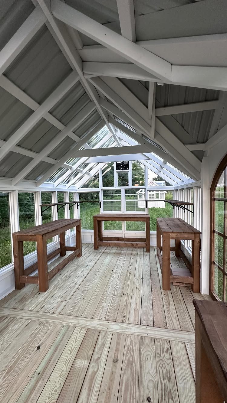 Interior of a white wooden greenhouse featuring three wooden work tables and glass windows.