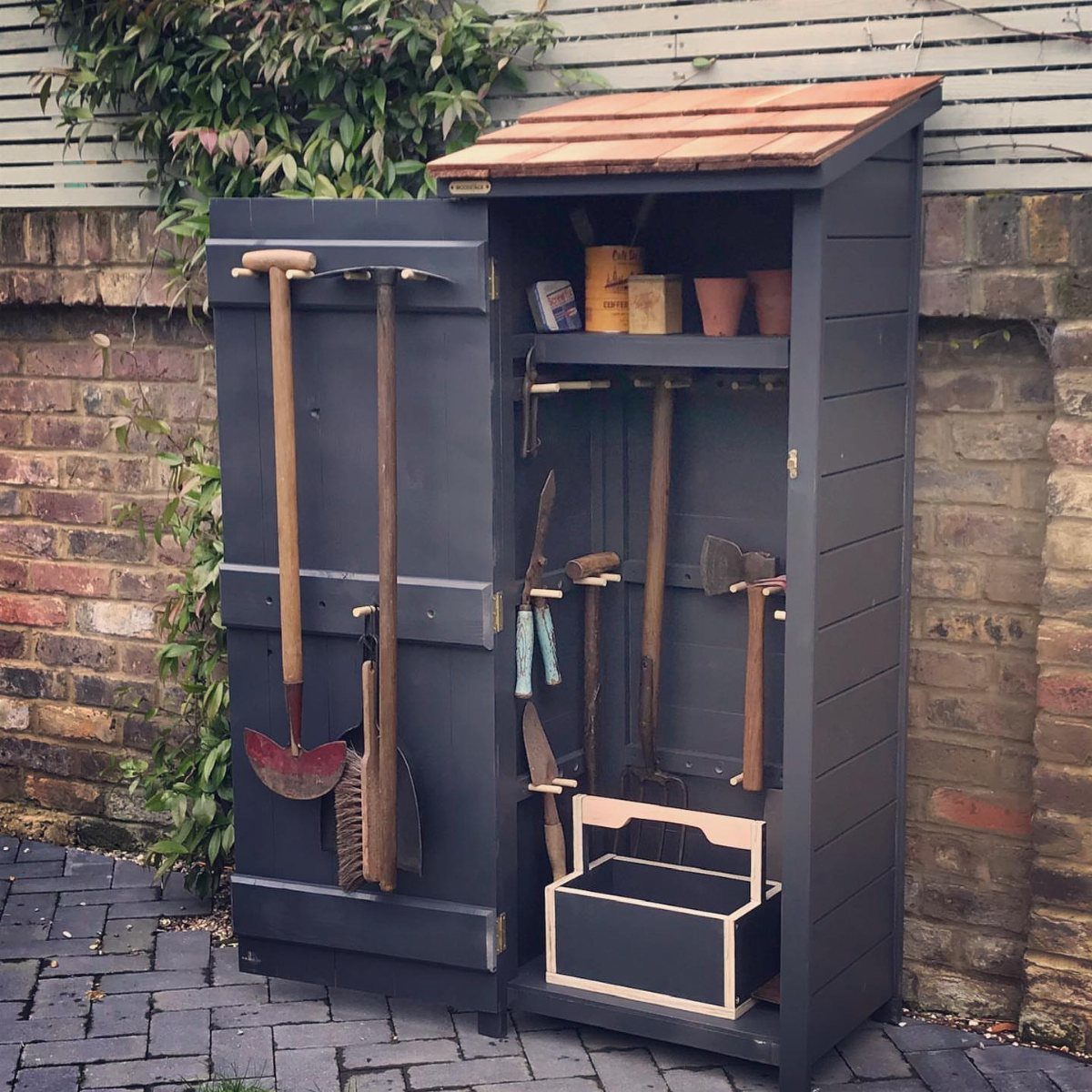 An open dark grey wooden mini garden tool shed displaying various handheld gardening tools and pots.