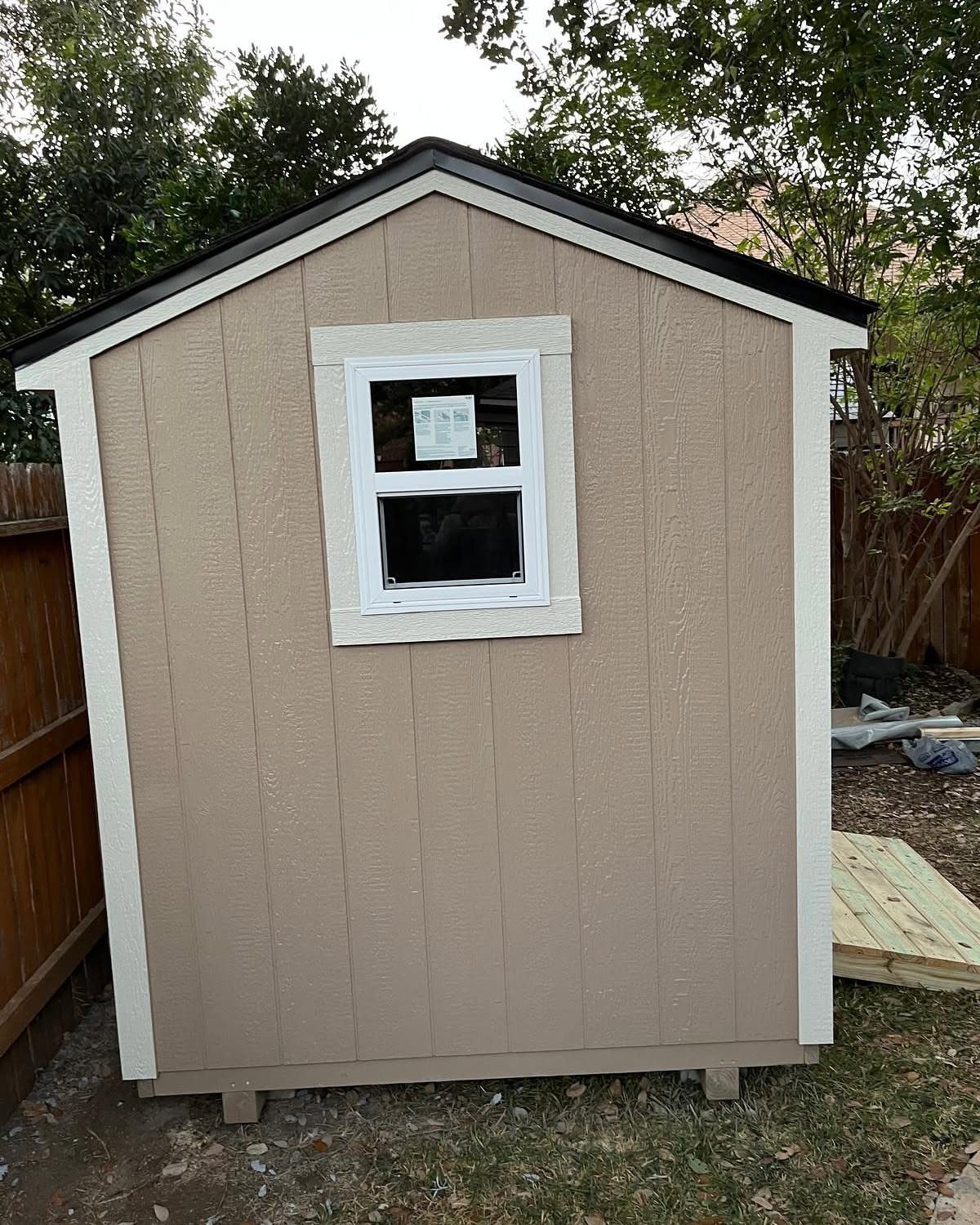 A small tan wooden storage shed with white trim and a single window outdoors.