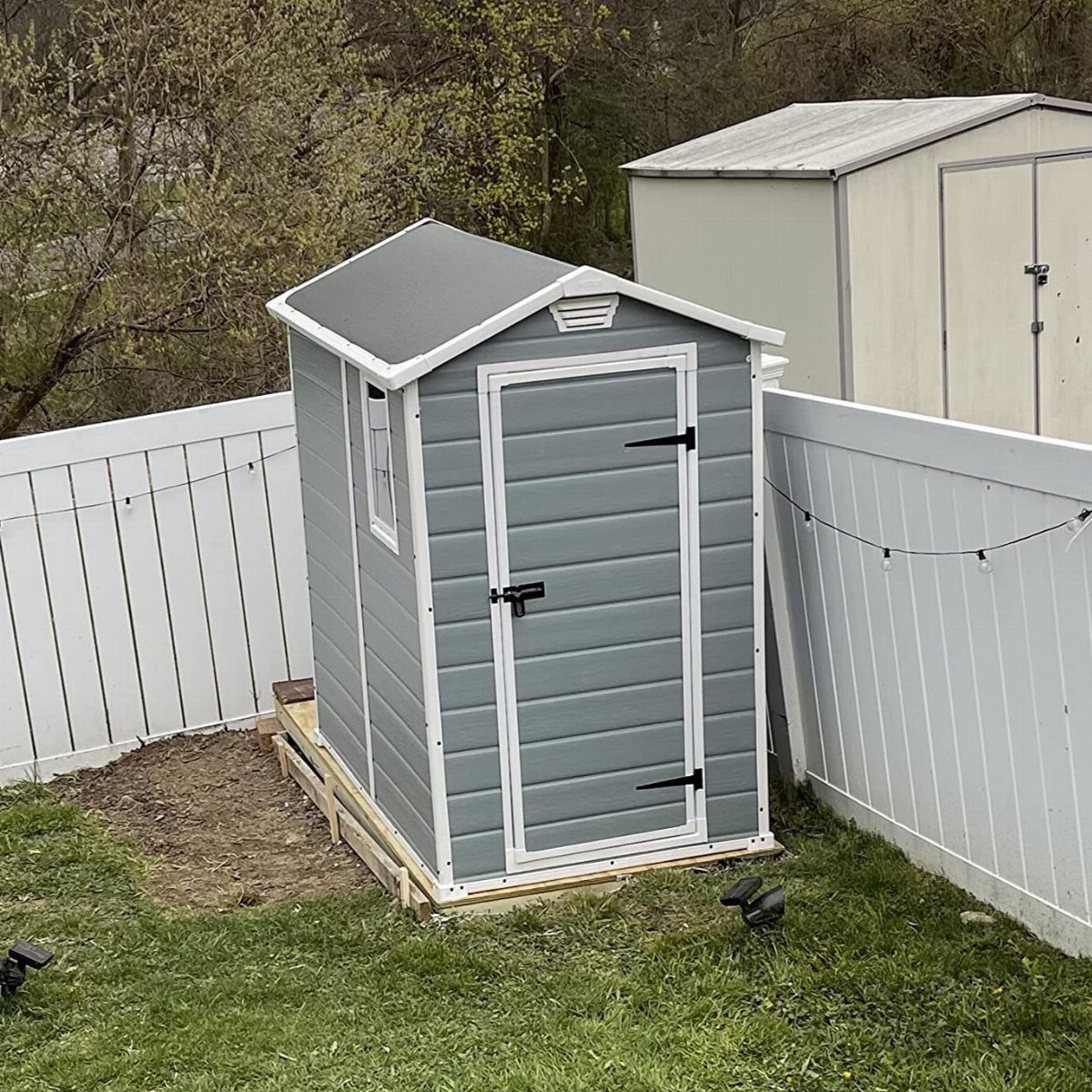 A small grey resin storage shed with white trim sits in a backyard against a white fence.