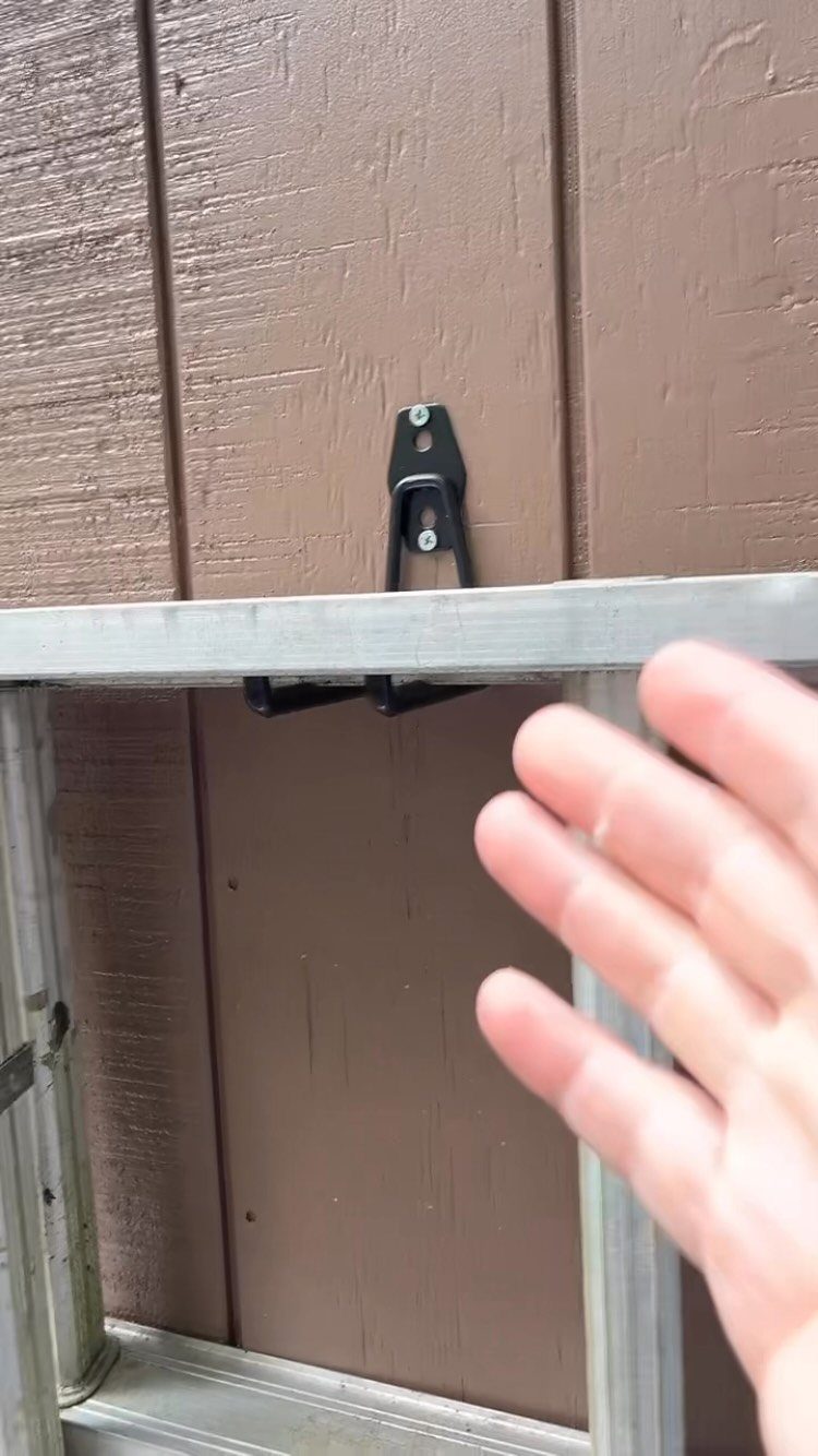 A black metal storage hook mounted on a brown wooden wall holding an aluminum ladder.