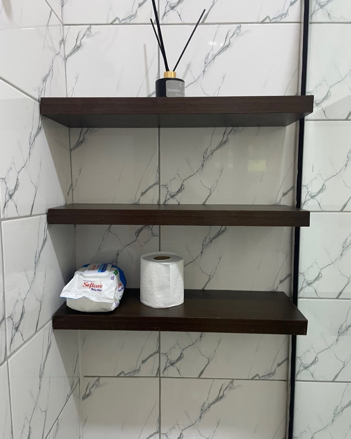 Three dark wood floating shelves mounted on white marble tiles holding bathroom essentials.