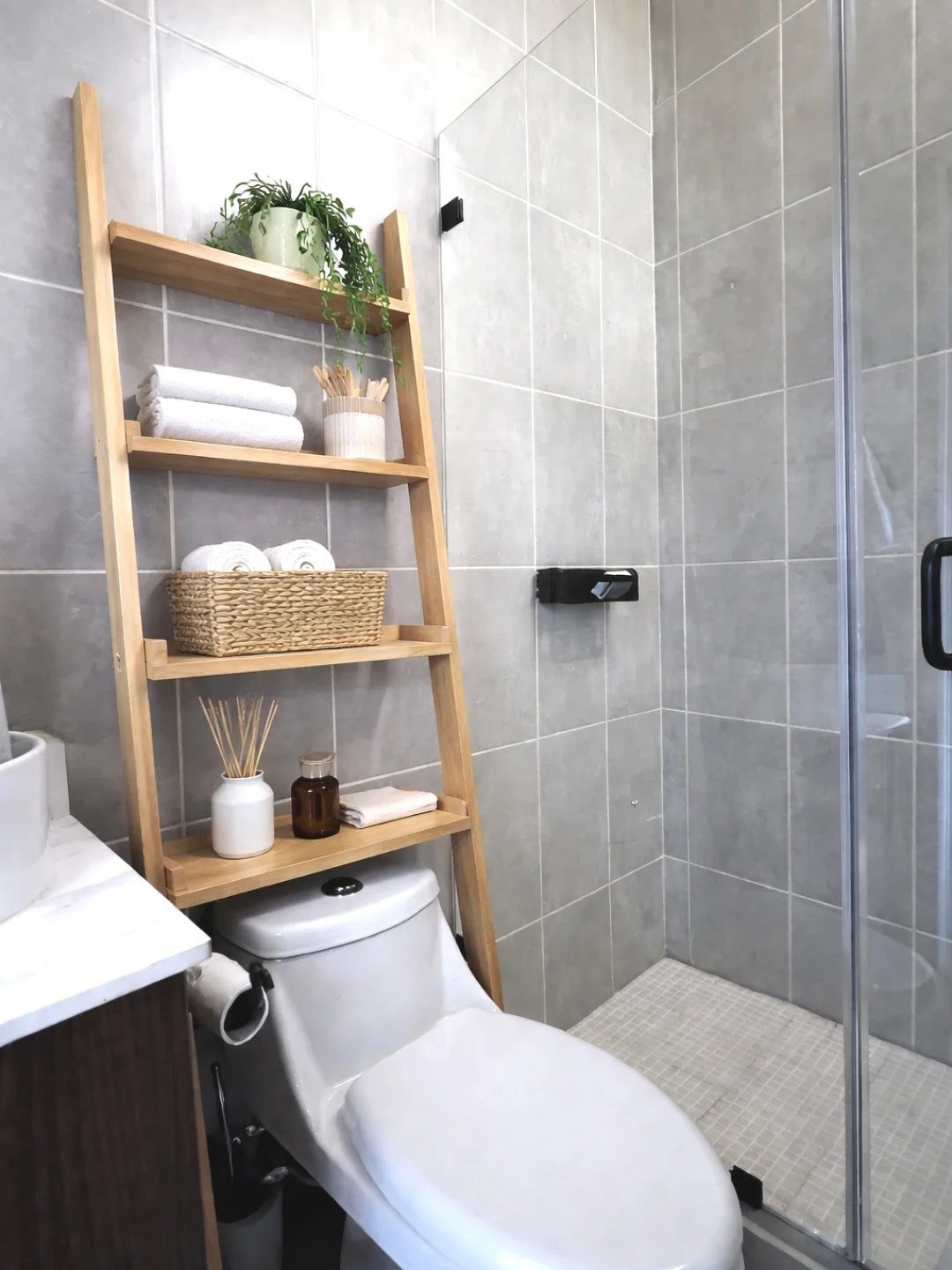 Wooden ladder shelves holding towels, a plant, and bathroom accessories against a grey tiled wall.