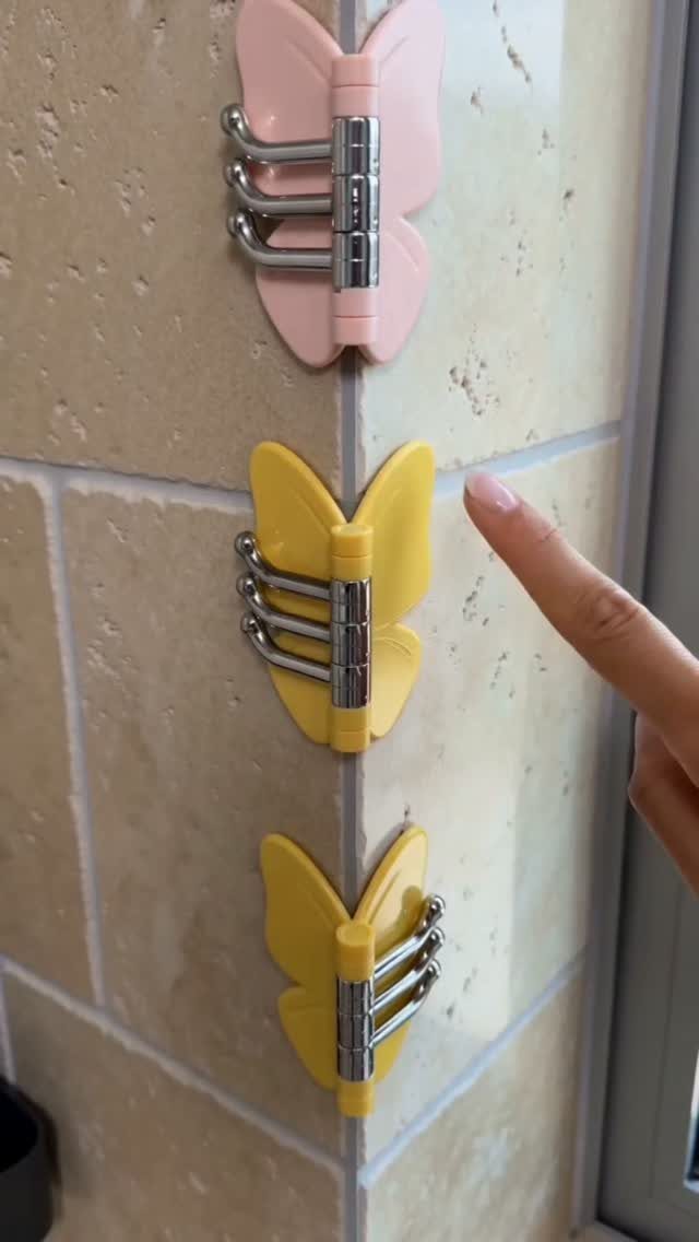 Three butterfly-shaped wall hooks in pink and yellow mounted on a tiled corner wall.