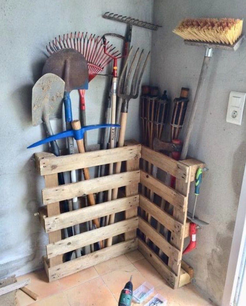 Garden tools stored upright in a corner organizer made from wooden pallets against a wall.