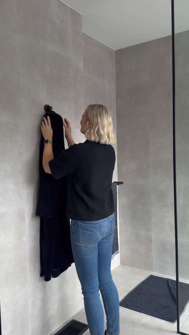 A woman hanging a black towel onto a wall-mounted hook in a grey tiled bathroom.