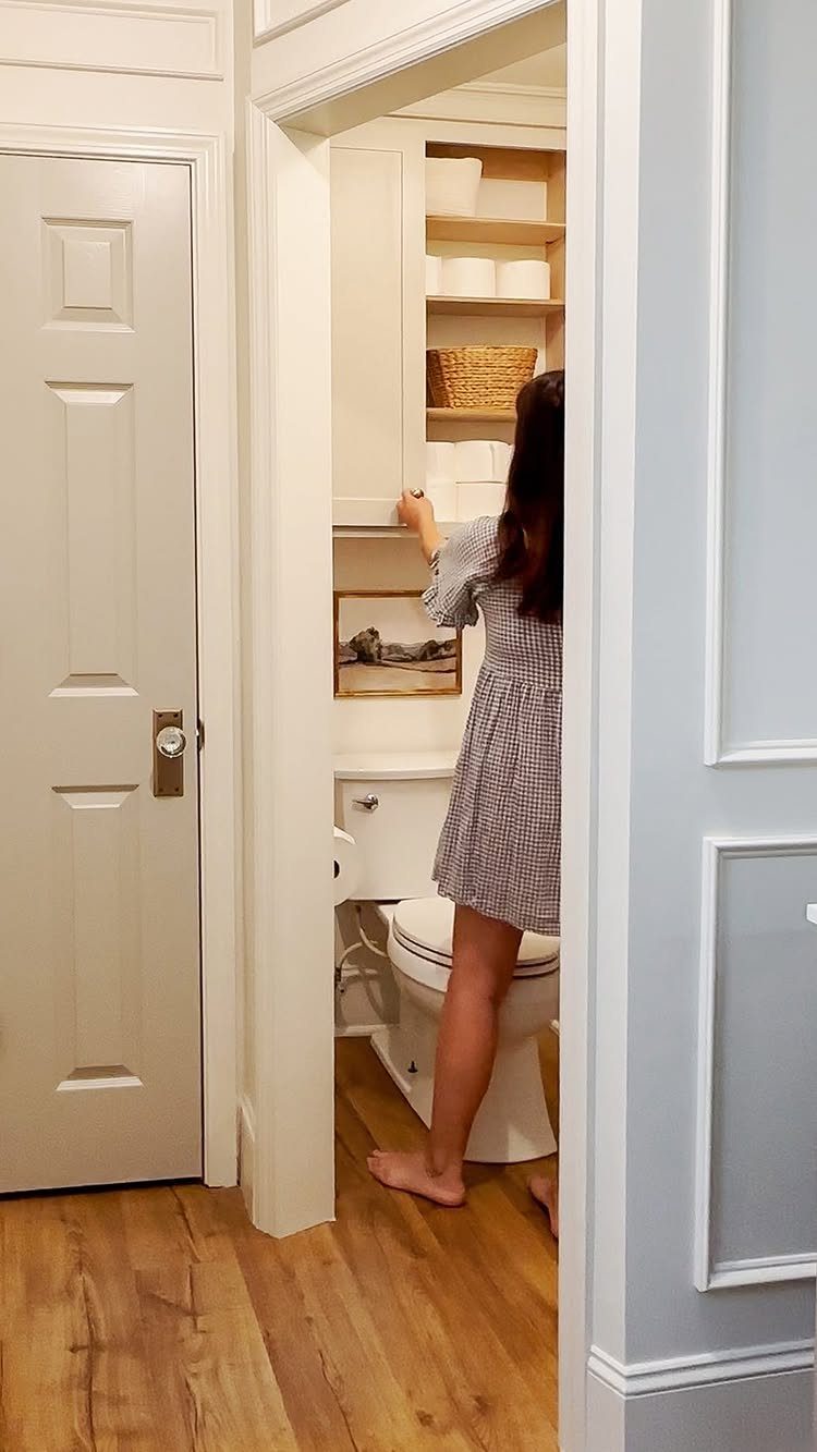 A person reaching for items on white built-in shelves located above a toilet in a bathroom.
